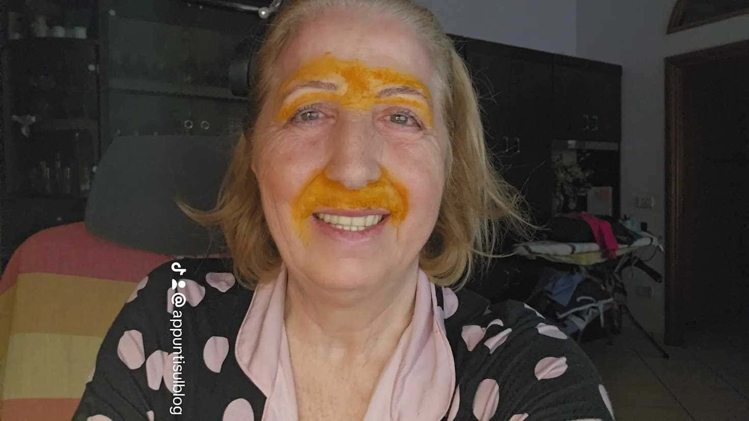 Ragazza sorridente con trucco originale di colore arancione sulla fronte e sul naso, in un ambiente domestico, ritratta mentre fa un selfie in casa.