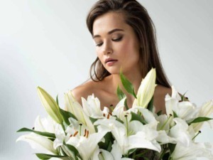 Bellissima donna con lunga chioma bruna che tiene un grande mazzo di gigli bianchi. Eleganza e fascino combinati con fiori freschi, ideale per articoli su bellezza, fiori e benessere.