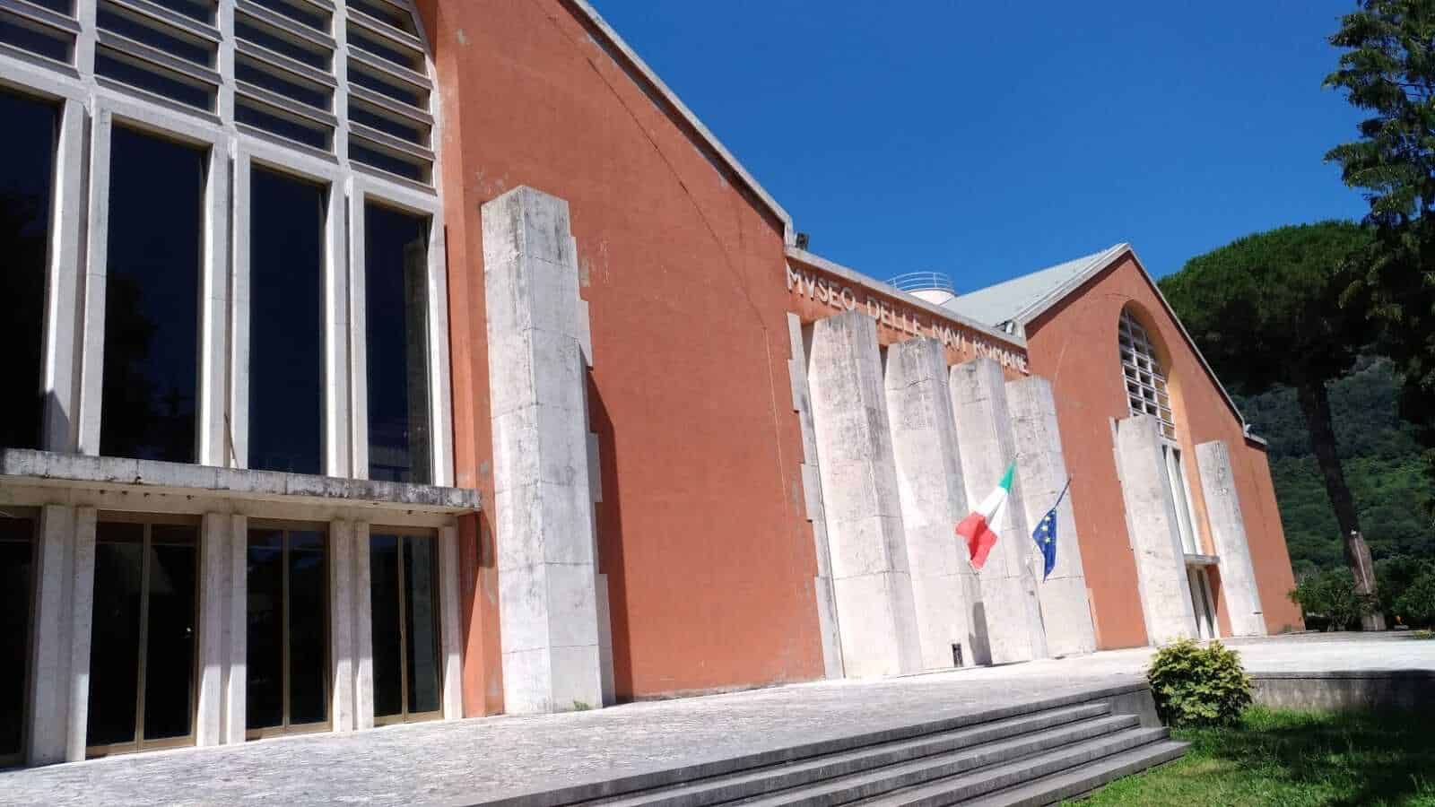 Museo delle Navi Romane di Nemi, edificio moderno con facciata in mattoni rossi e grandi finestre frontali, situato tra le colline verdi, sede delle esposizioni archeologiche e storiche sulle antiche navi romane.