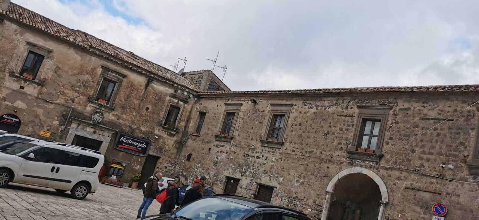 Antico edificio in pietra con finestre di legno, architettura storica italiana, piazza con auto e persone, atmosfera di paese mediterraneo, centro storico orientato a turismo e cultura.