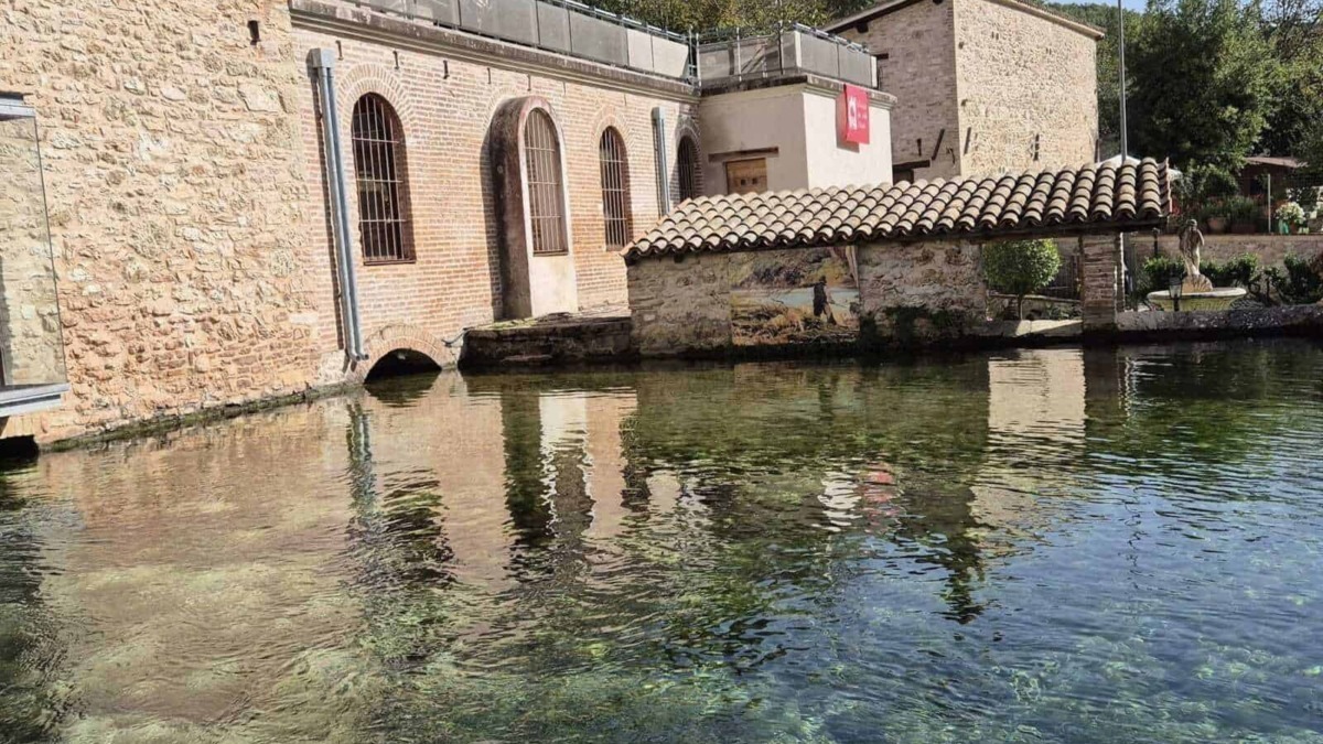 Vecchia centrale idroelettrica ristrutturata con pareti di mattoni e finestre ad arco, situata sul bordo di un lago naturale circondato da vegetazione.