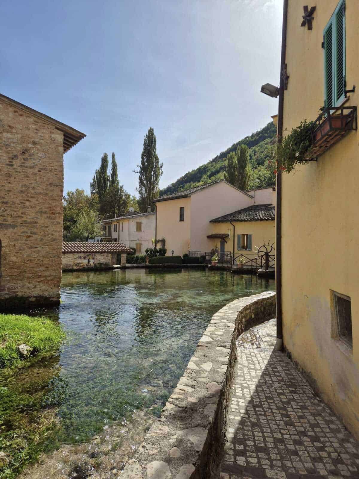 Vecchio paesino italiano con case colorate, mulino e affascinante fontana lungo il fiume, immerso nella natura delle colline, perfetto per relax e fotografia.