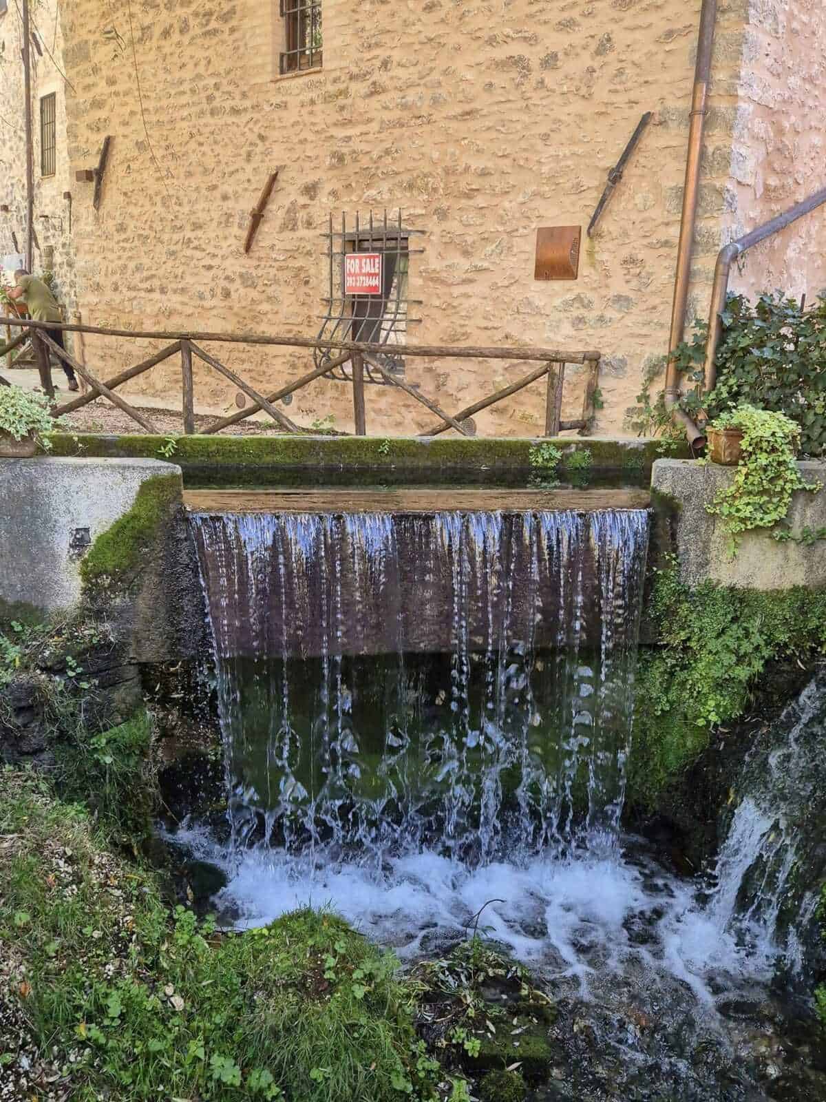 Arroyo urbano con cascata in pietra lungo un edificio storico in stile toscano, con vivaio di piante e balcone in legno, ambiente rustico e suggestivo per fotografie di paesaggi e natura.