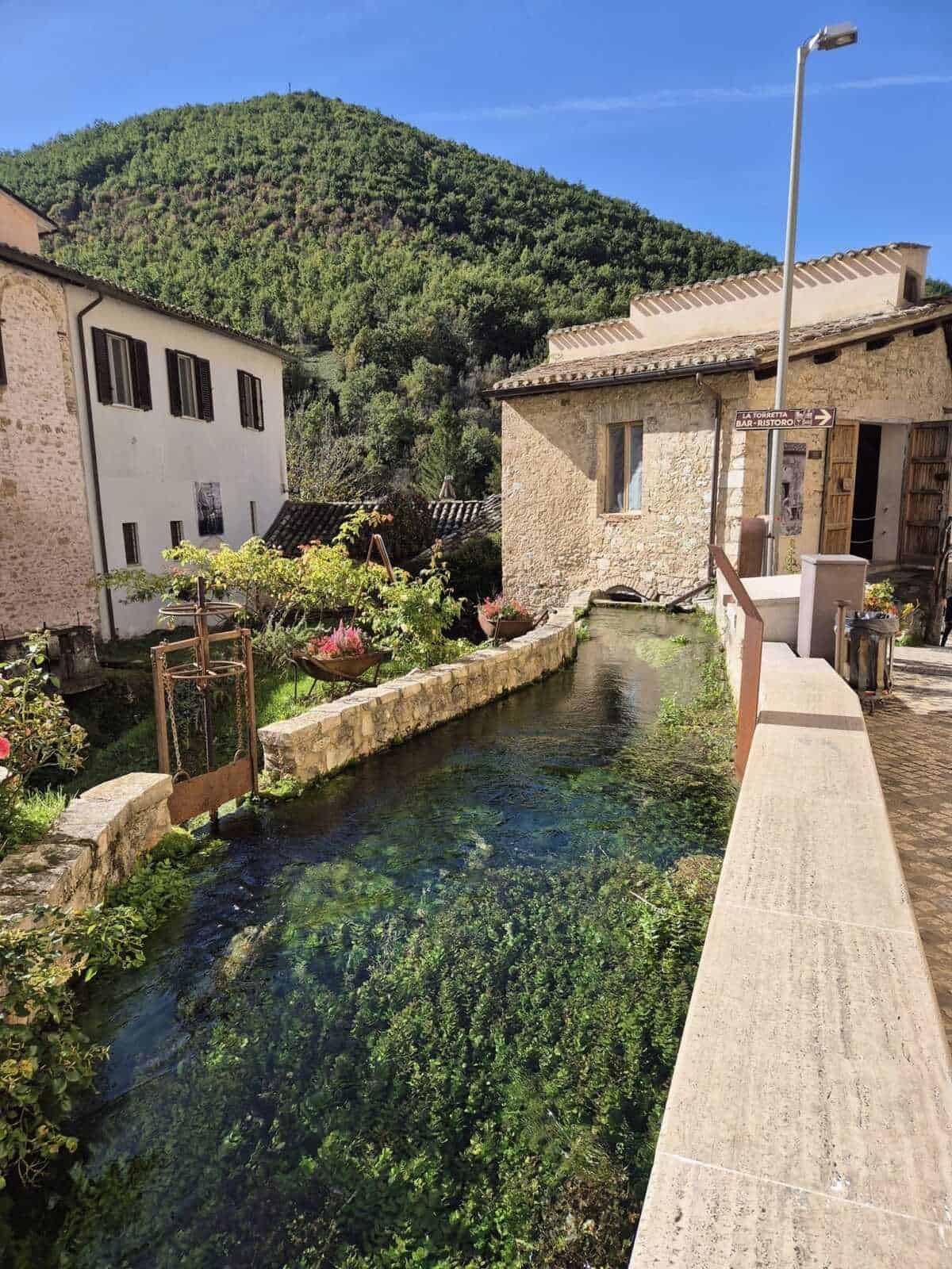 Fiume in un piccolo villaggio con case in pietra, fiori e una vista sulle colline verdi sotto un cielo azzurro, creando un'atmosfera italiana suggestiva e tranquilla.