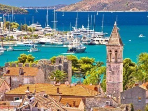 V junkyard, boats docked at marina in a coastal town with historic stone buildings and a prominent bell tower under sunny sky.