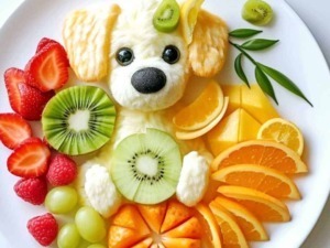 Colorato piatto di frutta con decorazione a forma di cane, con frutta fresca come kiwi, fragole, uva, arance e mandarini, e un mignolo di dolcezza.