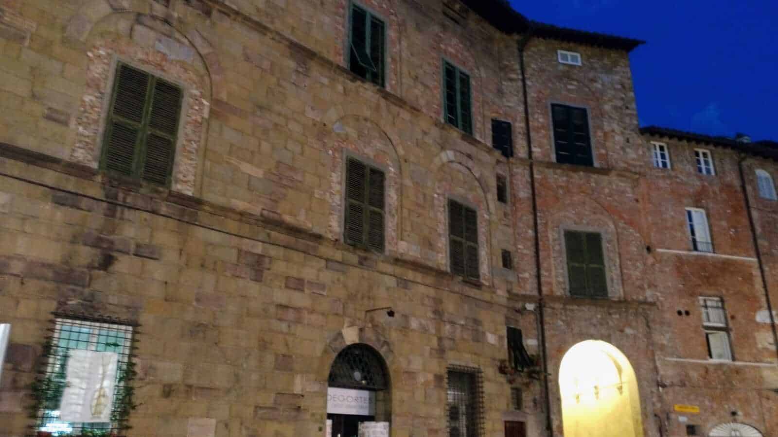 Antica facciata in pietra di un edificio storico al tramonto, con finestre con persiane verdi e arco illuminato, rappresenta l’architettura medievale toscana