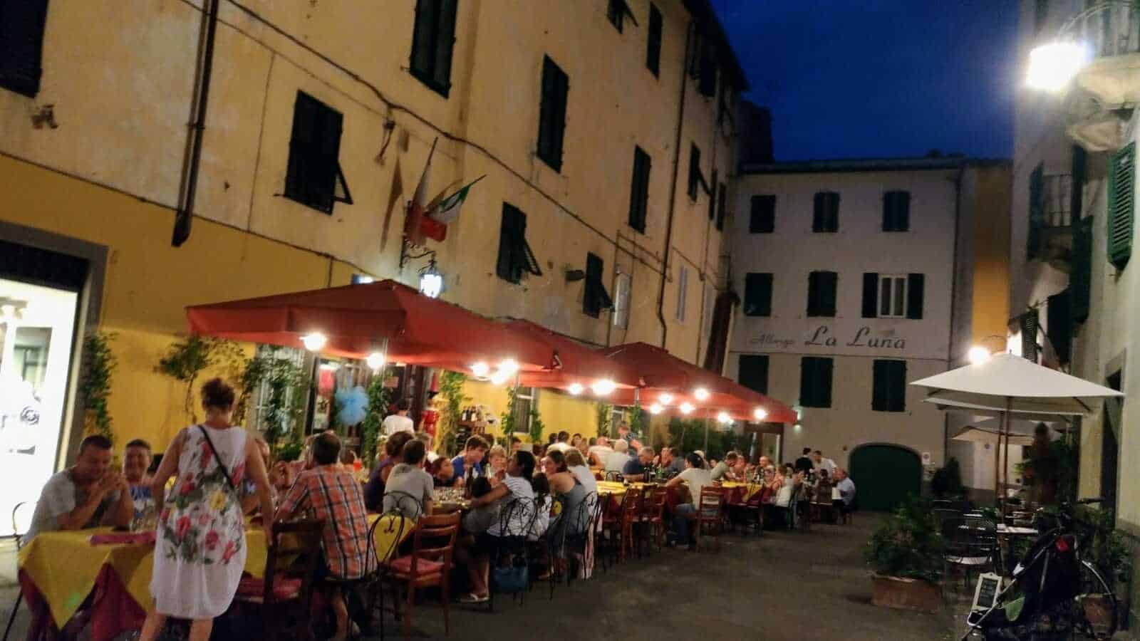 Ristorante all'aperto in centro storico durante la sera con tavoli, luci calde e clienti che cenano all'aperto, atmosfera vivace e accogliente.