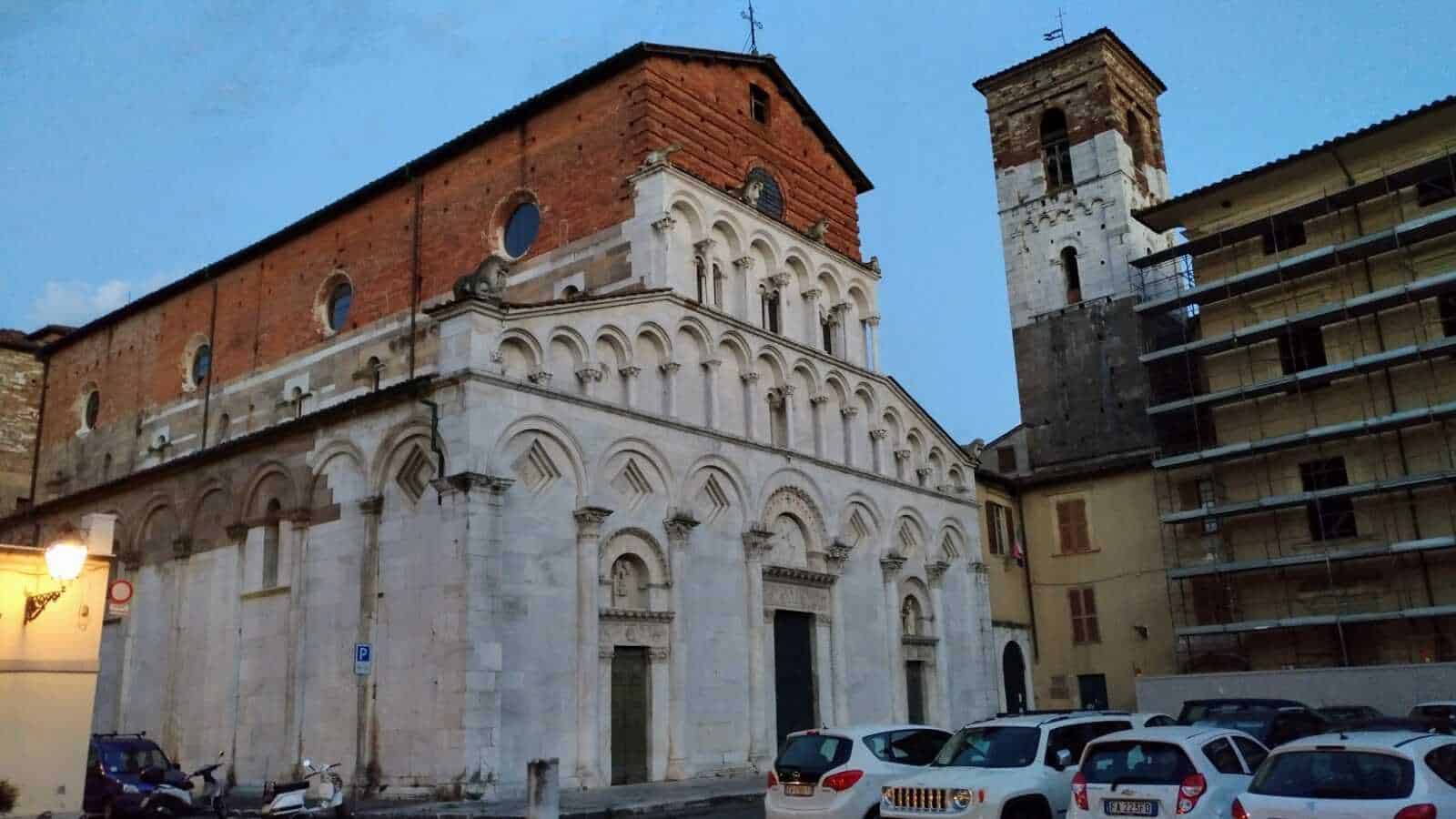 Vecchia chiesa storica con facciata in marmo e campanile in pietra, immerse nel centro di un borgo italiano con auto parcheggiate e edifici circostanti.