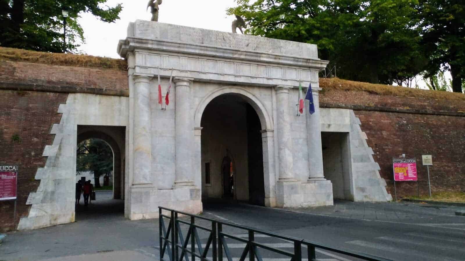 Antico ingresso monumentale con archi in marmo bianco, targhe e bandiere italiane e europee, sito storico nel centro di Lucca, Italia, immerso nel verde e in stile classico.