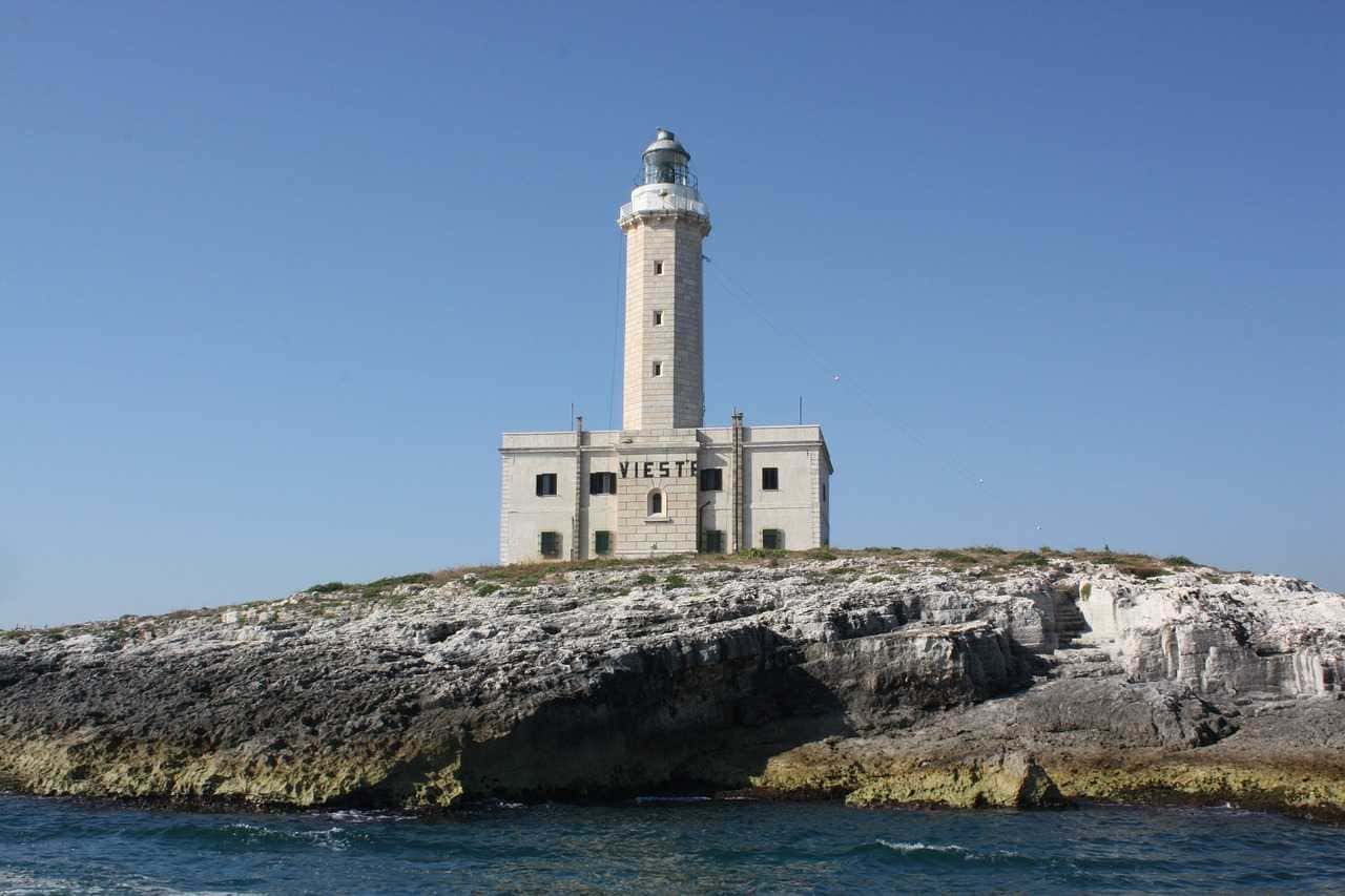 È il momento giusto per prenotare le tue vacanze estive 3 Faro di Vieste situato sulla scogliera a picco sul mare, simbolo del promontorio di Vieste, in Puglia, Italia, con cielo azzurro e acque cristalline.