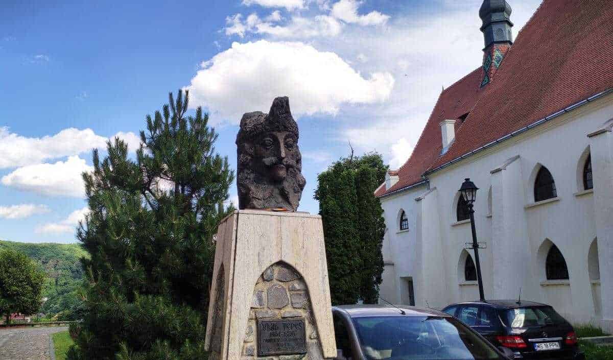 Statua storica e monumento nel paesaggio di un centro storico con edificio religioso e auto in primo piano, in un contesto di natura e architettura tradizionale.