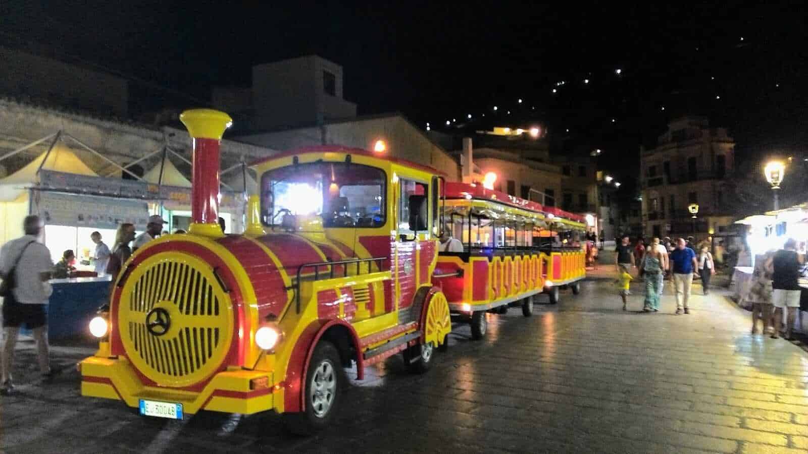 Colorato trenino turistico a trazione elettrica per città, perfetto per visite guidate e gite notturne, circondato da persone e negozi vivaci, in una piazza affollata e illuminata.
