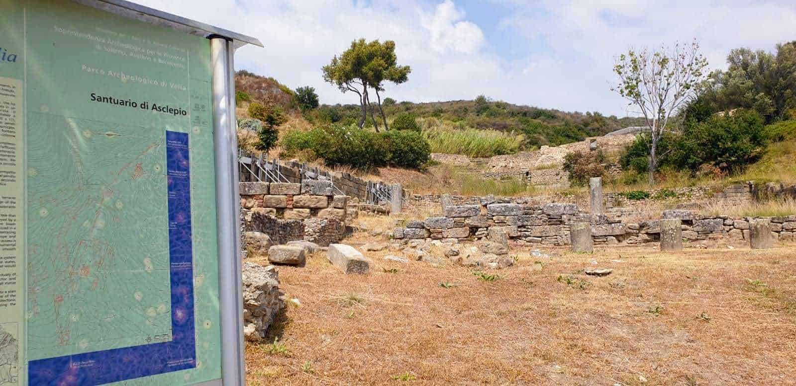 Resti archeologici dell'antico santuario di Asclepio in un sito immerso nella natura, con pannello informativo e paesaggio collinare circostante. Ideale per appassionati di archeologia e storia antica.