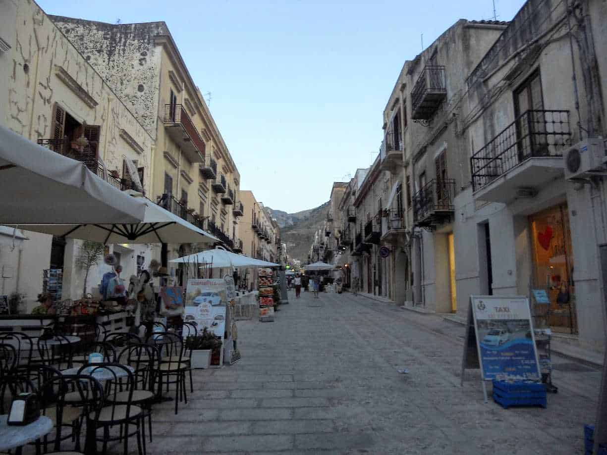 Vicolo pittoresco con caffè all'aperto e negozi in un borgo storico italiano con edifici antichi e montagne sullo sfondo.