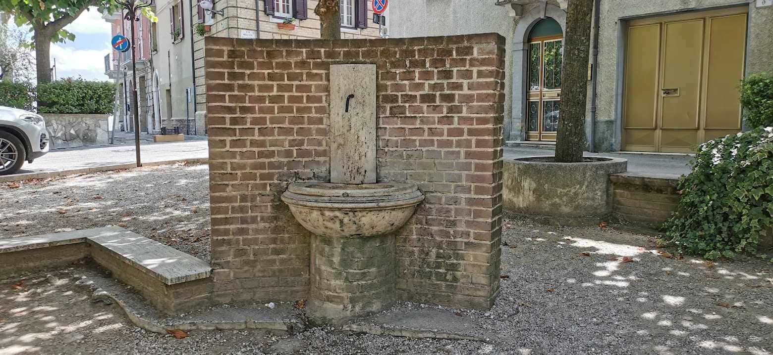 Fonte pubblica d'acqua in pietra e mattoni in un cortile urbano, circondata da edifici storici e strade, offre un angolo di tranquillità e storia nel quartiere.