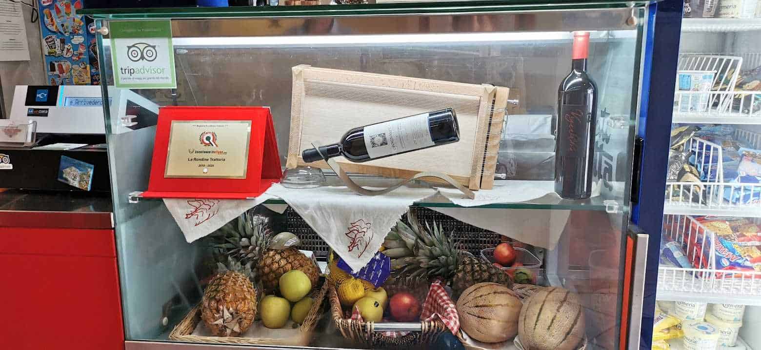 Vetrina frigo con frutta, vino, premio e slogan di TripAdvisor, in un negozio alimentare italiano.