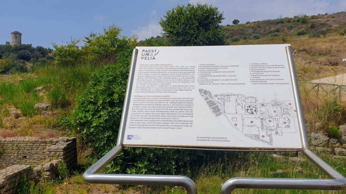 Cláudio di un pannello informativo all'aperto su antica archeologia e tradizioni della regione Paestum e Velia, situato in un'area naturale con colline e rovine storiche sullo sfondo.