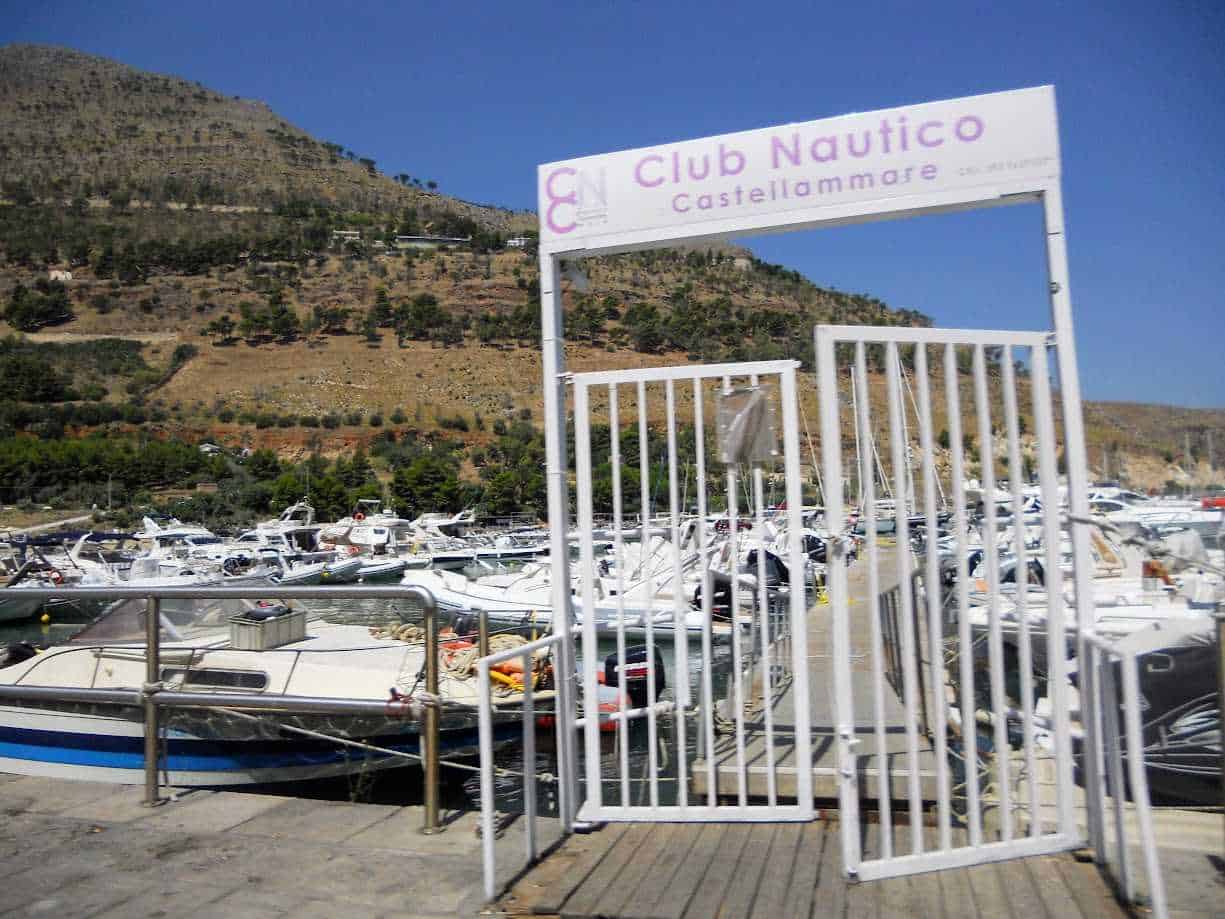 Cn Club Nautico Castellammare del Golfo, porto turistico con barche e yacht, ingresso con cancellata bianca, paesaggio montano sullo sfondo, cielo sereno, immagine di arredo marittimo, vacanze e nautica.