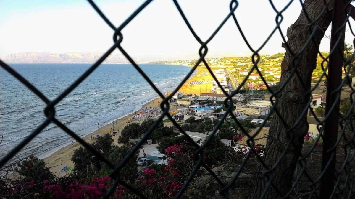 Frequenza di rete vista attraverso una recinzione, con spiaggia, mare, case e colline sullo sfondo, scena costiera italiana con atmosfera estiva e paesaggi marittimi affascinanti.