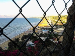 Frequenza di rete vista attraverso una recinzione, con spiaggia, mare, case e colline sullo sfondo, scena costiera italiana con atmosfera estiva e paesaggi marittimi affascinanti.
