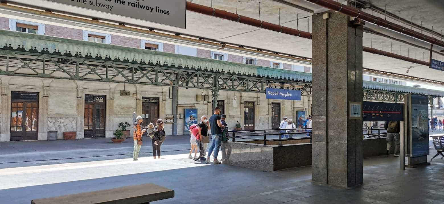 Stazione Napoli Mergellina, persone in attesa del treno sulla piattaforma ferroviaria, atmosfera urbana, viaggiatori con bagagli, dettagli architettonici, ambiente di trasporto pubblico.