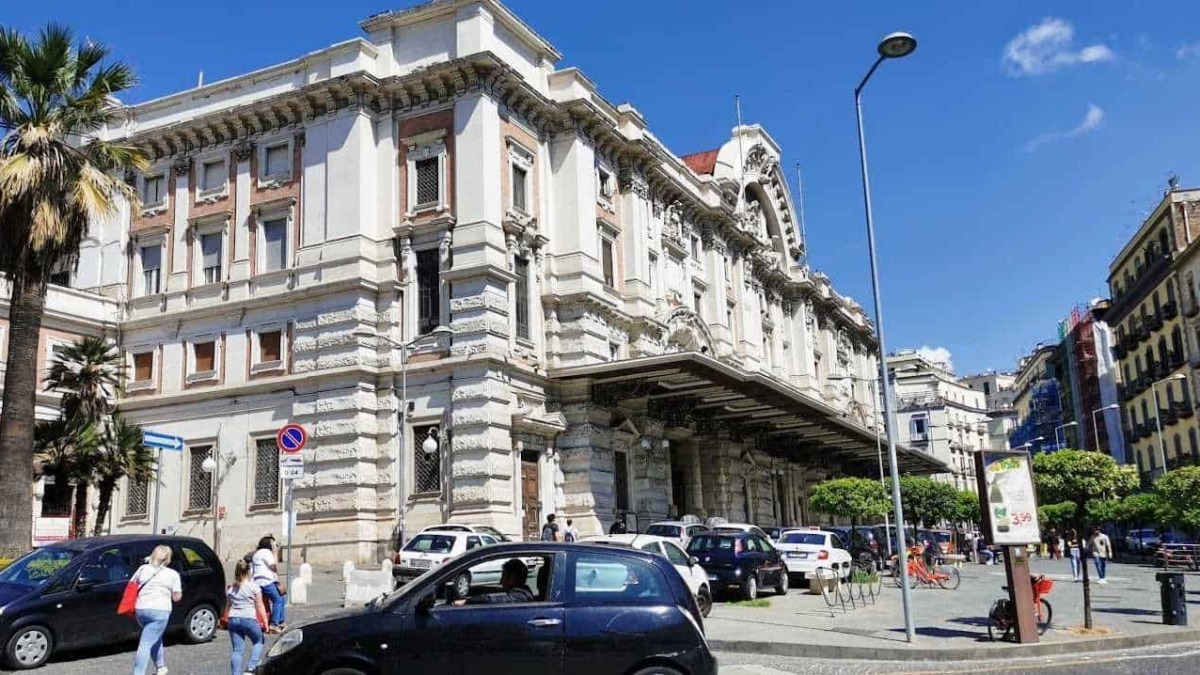 Palazzo storico sulla strada principale di Napoli, con architettura elegante e dettagli decorativi. Strada trafficata con auto, bici e pedoni sotto un cielo azzurro.