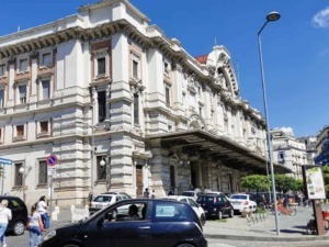Palazzo storico sulla strada principale di Napoli, con architettura elegante e dettagli decorativi. Strada trafficata con auto, bici e pedoni sotto un cielo azzurro.