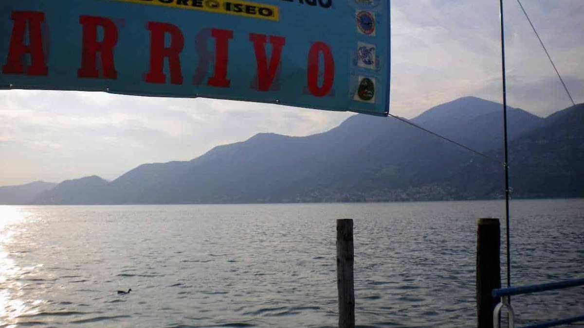 Passeggiata sul Lago di Monate con vista sulle montagne, banner che annuncia la XIII traversata del lago, attività estive nel predore e Iseo, spazi aperti e natura in Lombardia.