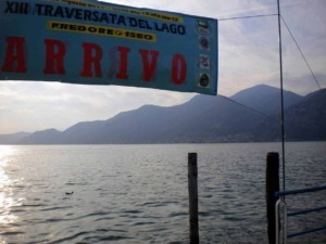 Passeggiata sul Lago di Monate con vista sulle montagne, banner che annuncia la XIII traversata del lago, attività estive nel predore e Iseo, spazi aperti e natura in Lombardia.