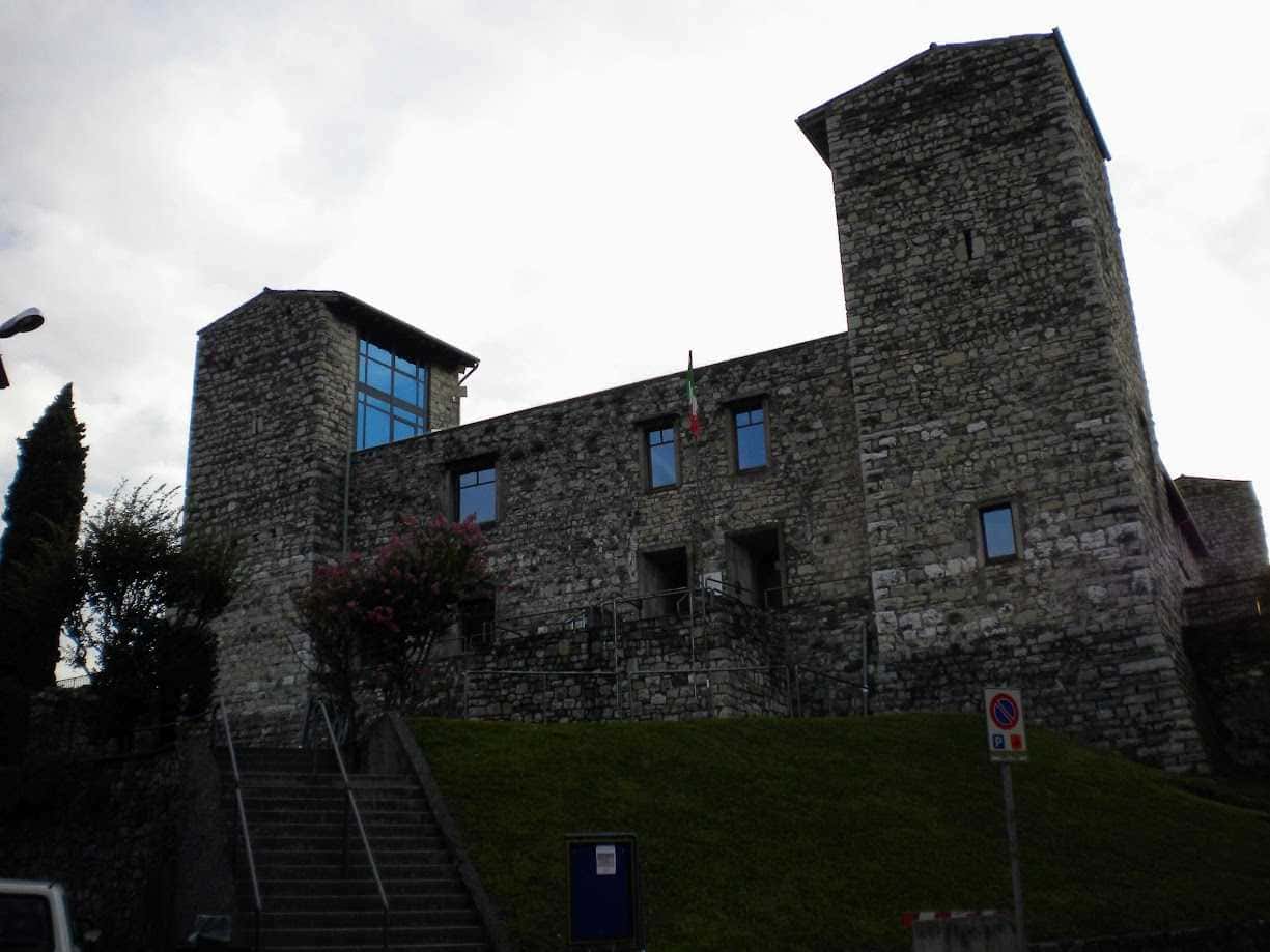 Cosa Vedere a Iseo città sul Lago di Iseo: centro storico, castello e lungolago 4 Castello Oldofredi, medievale in pietra con torri, situato su una collina, con bandiera italiana, palazzi storici, vista panoramica, in Italia, esempio di architettura storica e patrimonio culturale.