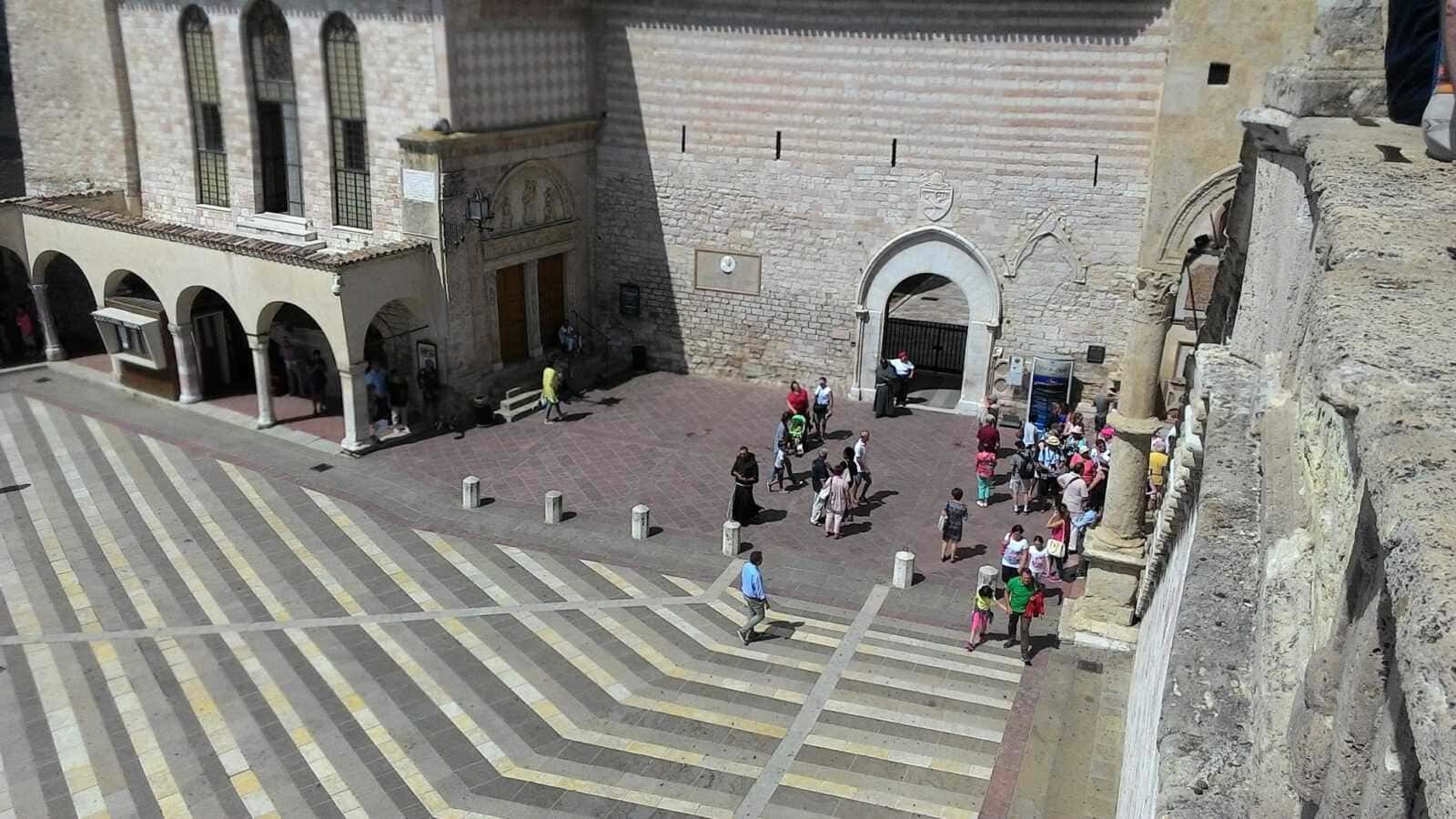 Piazza storica di Assisi con persone, architettura antica e pavimentazione decorativa, scena urbana nel centro di una città italiana