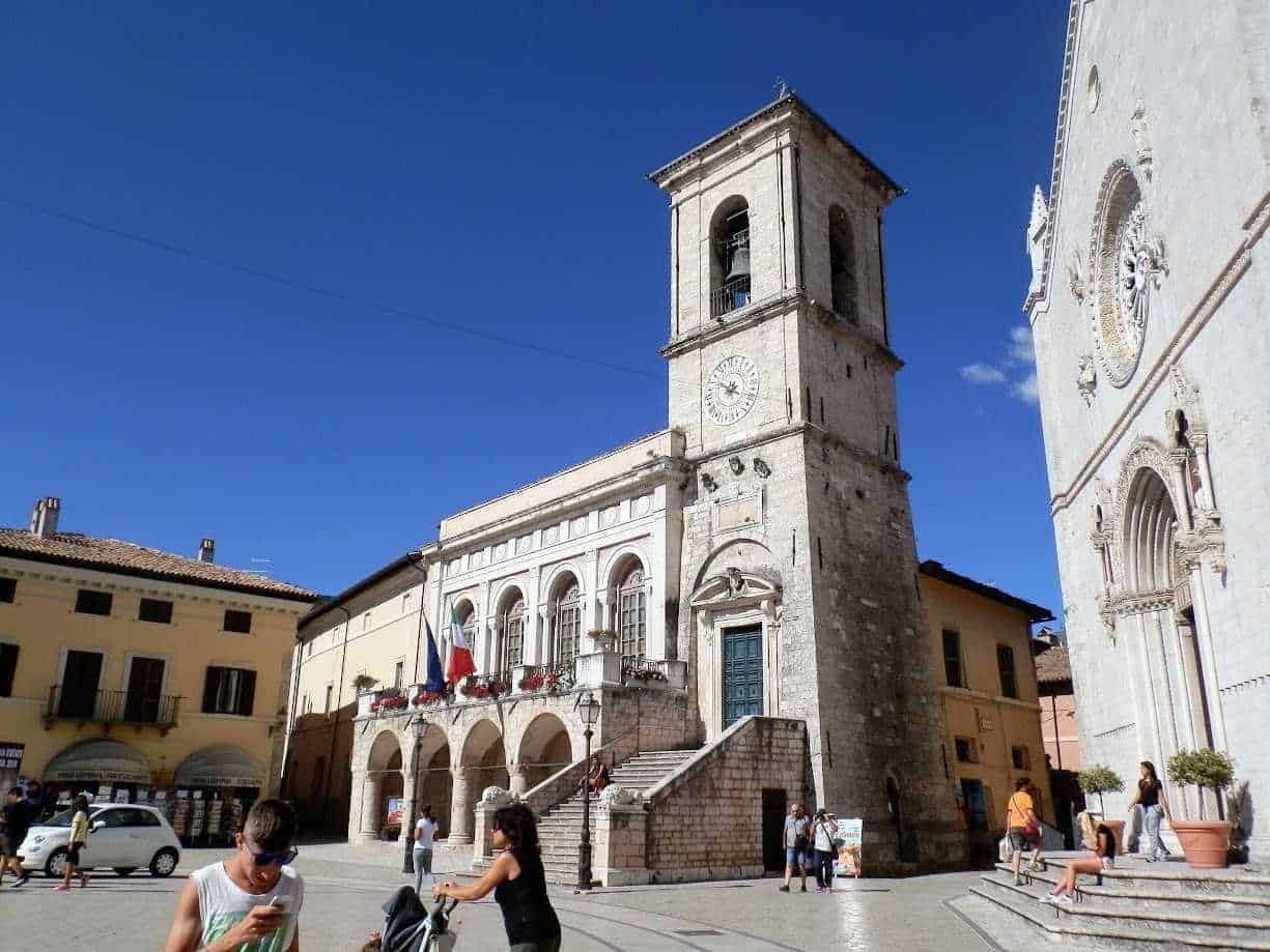 Campanile storico in piazza con persone e auto, edifici antichi con facciata decorata, cielo blu e giornata di sole, atmosfera vivace nel centro di un borgo italiano.