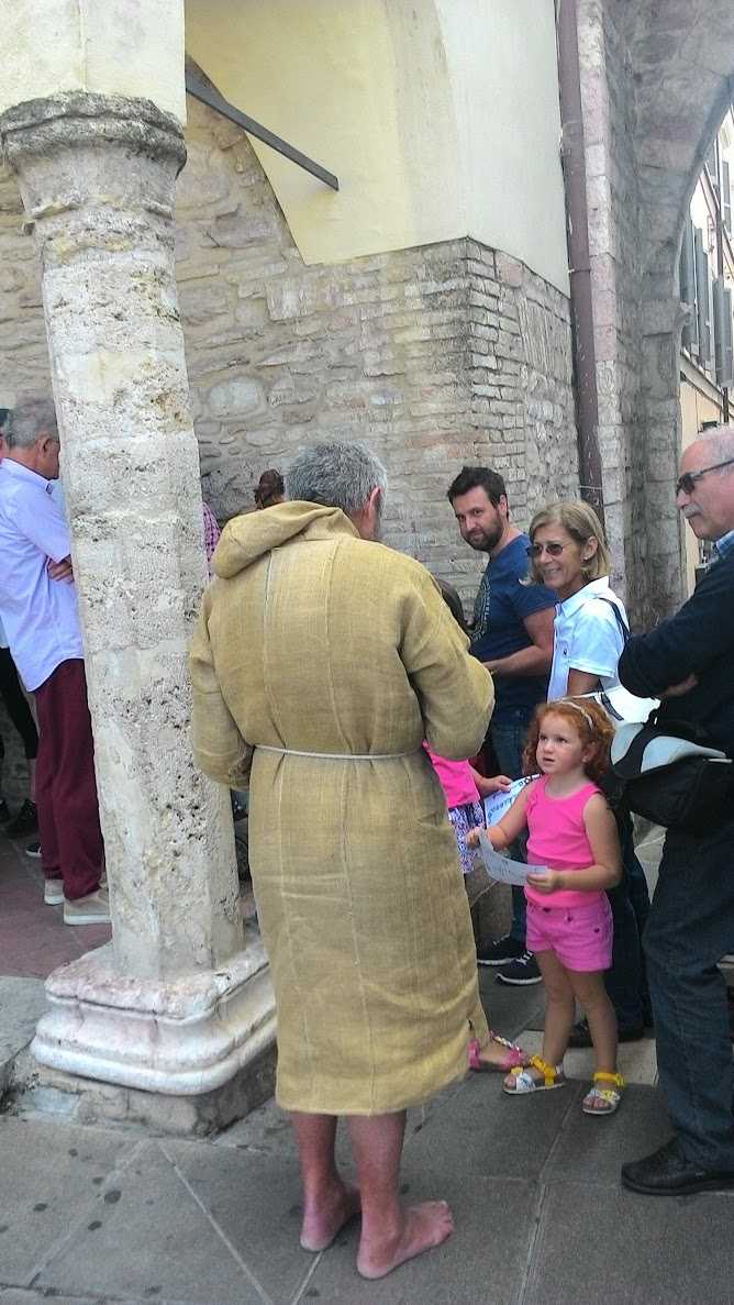 Piazza storica di Assisi con persone che chiacchierano vicino a colonne in pietra e mura antiche