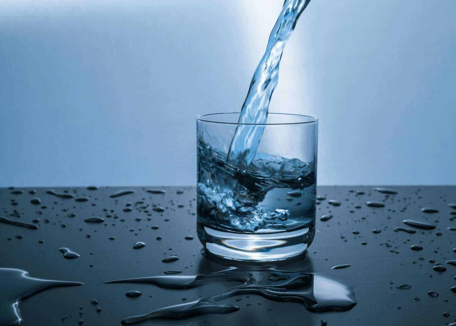 Acqua che viene versata in un bicchiere trasparente con gocce e schizzi sparse sulla superficie nera, evidenziando purezza e freschezza.