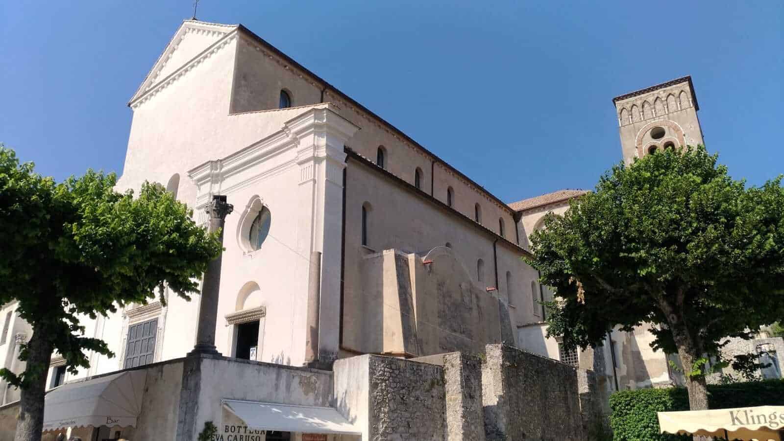 Antico edificio religioso con torri e facciata bianca, circondato da alberi verdi e situato in un centro storico soleggiato.