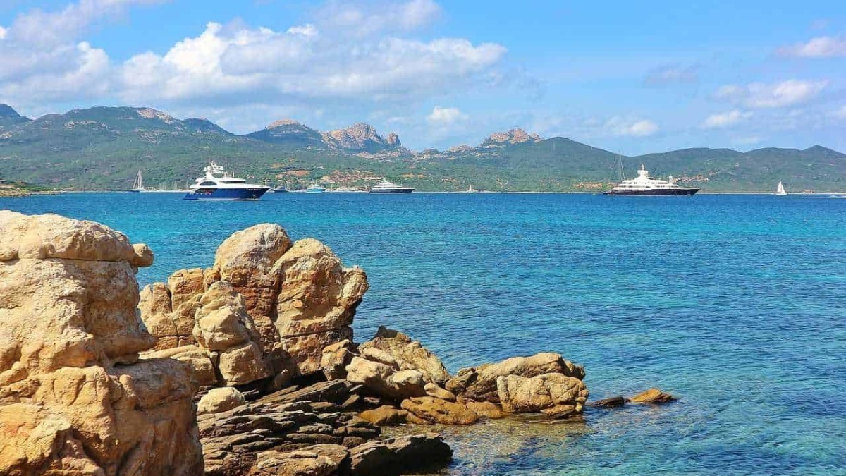 Baia con yacht di lusso e montagne sullo sfondo, riviera mediterranea, mare cristallino, coste rocciose, vacanze estive in Italia, paesaggi marini, turismo di lusso.