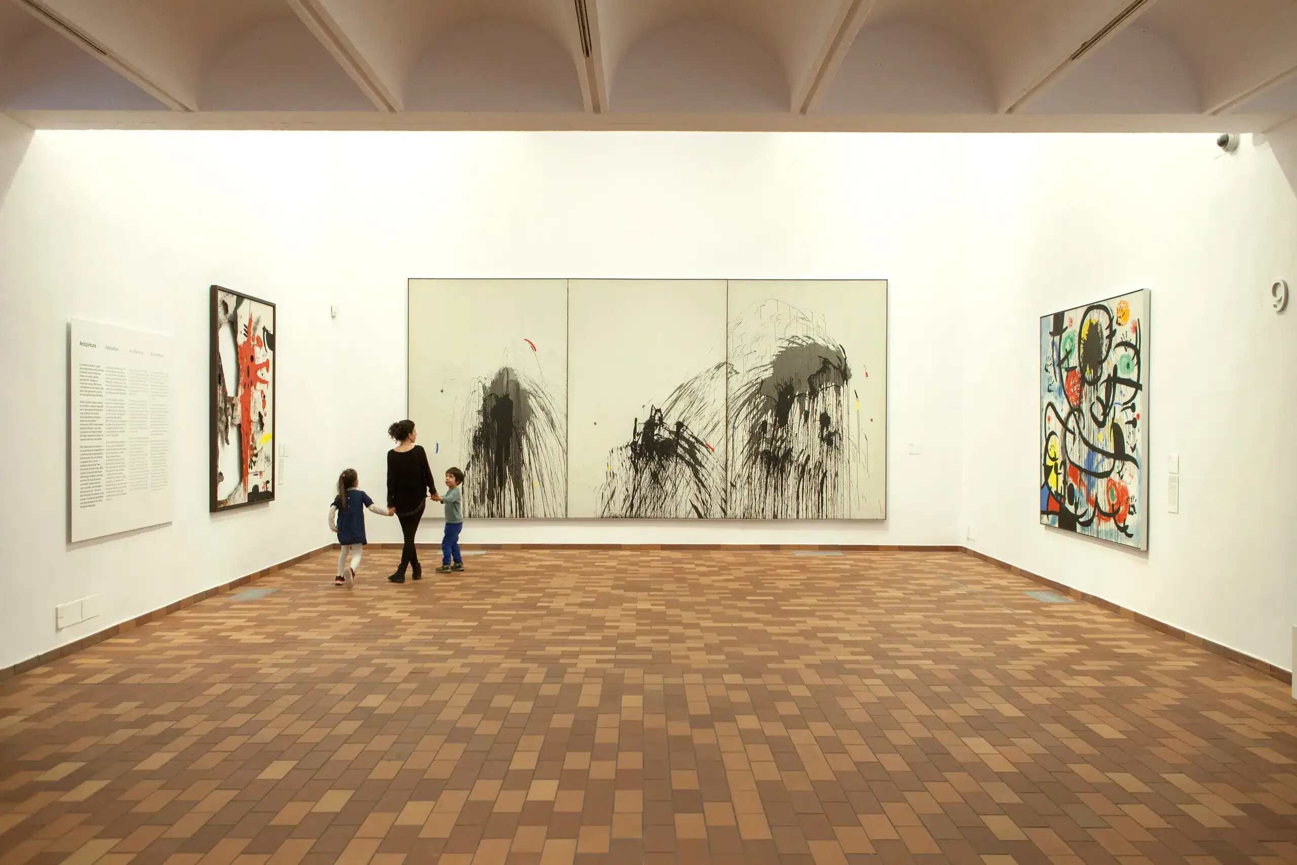 Dipinto astratto bianco e nero in una galleria d'arte, con due bambini e un adulto che osservano l'opera, pavimento in piastrelle di mattone, pareti bianche e illuminazione naturale.