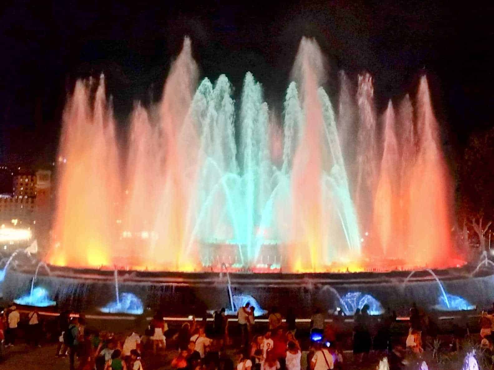 Faro luminoso con giochi d'acqua e luci colorate, spettacolo serale in una piazza, atmosfera festiva, fontane danzanti illuminate, momento di intrattenimento per famiglie e turisti.
