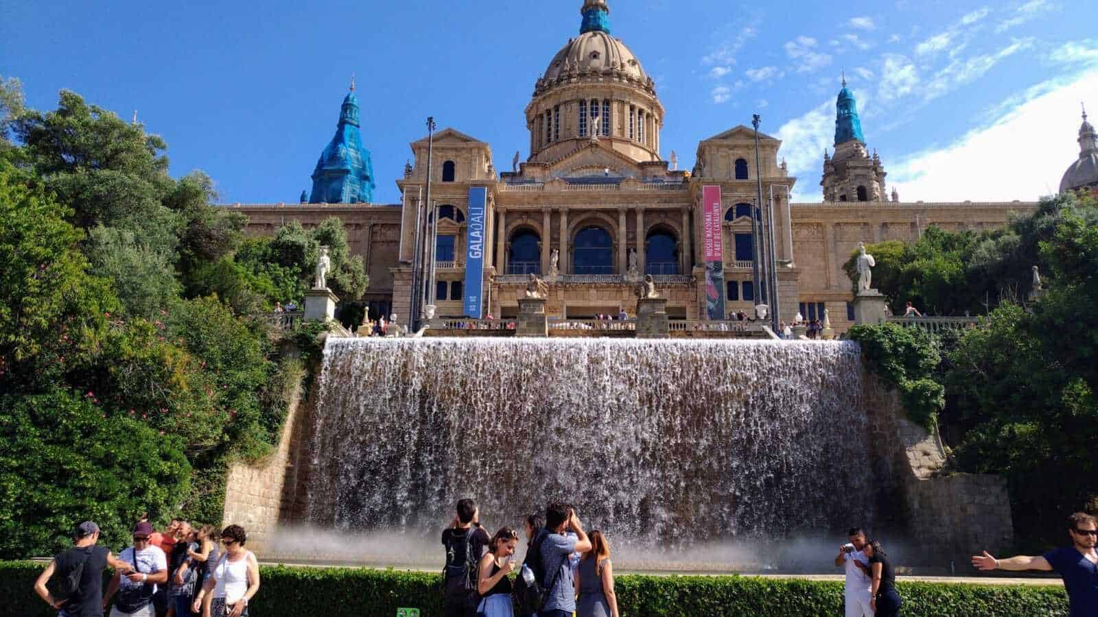 Montjuïc a barcellona: Vista panoramica del Palau Nacional e della collina di Montjuïc, con cascata davanti e turisti che ammirano l'architettura storica in una splendida giornata di sole.