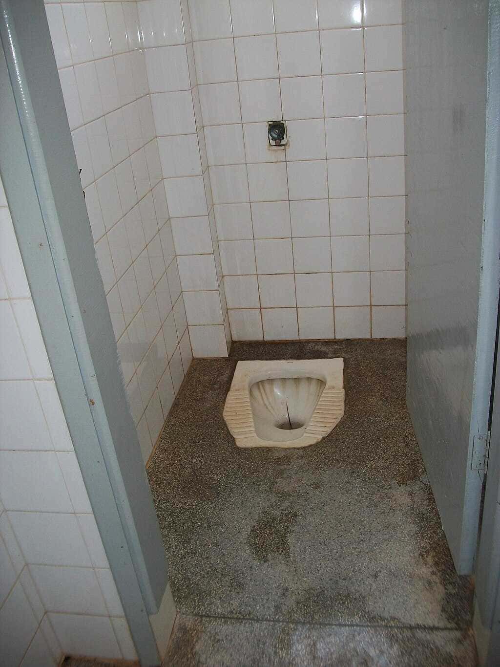 Scarico pertoilettes tradizionale in bagno pubblico con piastrelle bianche e pavimento in cemento, senza sedile. Ambiente usato o in fase di ristrutturazione.