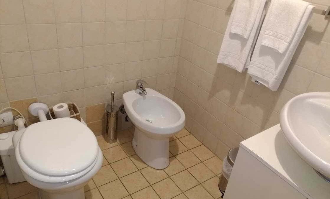 Spazio bagno con wc, bidet, lavabo e asciugamani bianchi, piastrelle beige per bagno funzionale e pratico. Ideale per luoghi di uso quotidiano con accessori essenziali e arredo semplice.