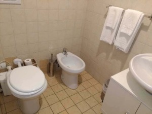 Spazio bagno con wc, bidet, lavabo e asciugamani bianchi, piastrelle beige per bagno funzionale e pratico. Ideale per luoghi di uso quotidiano con accessori essenziali e arredo semplice.