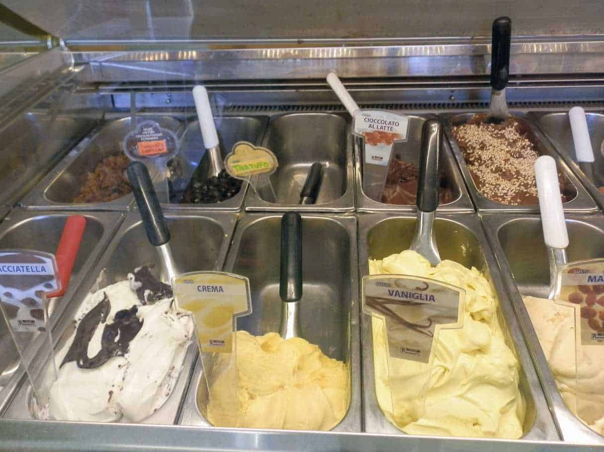 Ampio banco di gelato artigianale con gusti come crema, vaniglia, cioccolato al latte e altre varietà, servito in una gelateria italiana. Disponibili anche gusti con topping e aromi variegati.