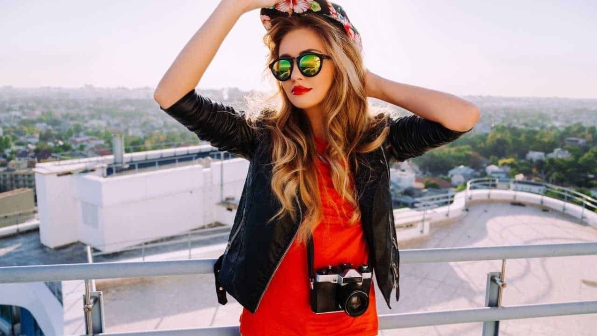 Ragazza con occhiali da sole, cappello floreale e fotocamera sul tetto di un edificio con vista sulla città, stile urban e moda estiva, perfetta per blog di lifestyle e viaggi.