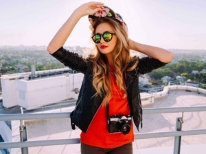 Ragazza con occhiali da sole, cappello floreale e fotocamera sul tetto di un edificio con vista sulla città, stile urban e moda estiva, perfetta per blog di lifestyle e viaggi.