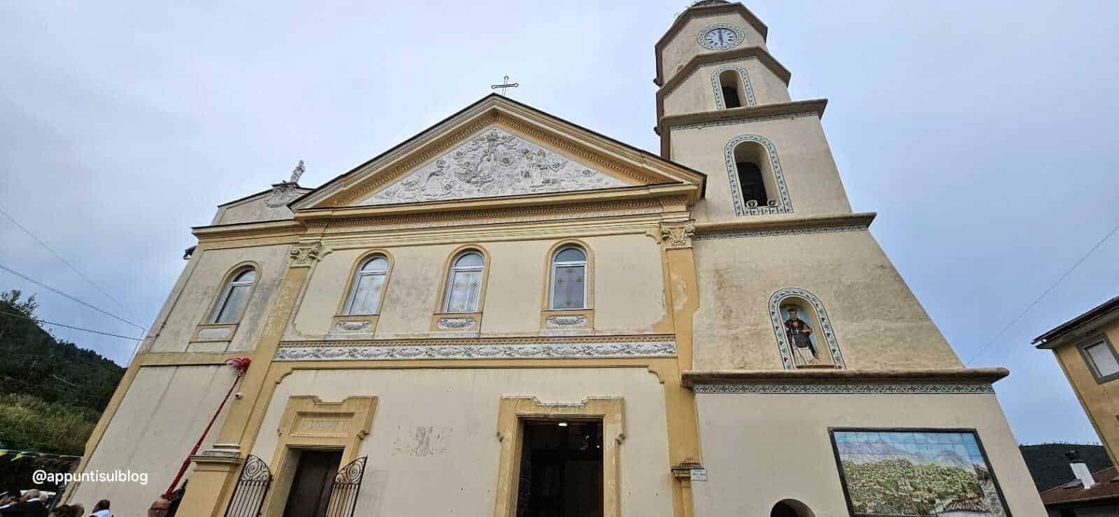 Antico municipio di montagna con scuola cattolica, architettura storica e campanile alto in pietra, dettagli decorativi e affreschi esterni, situato in un pittoresco paesaggio collinare.