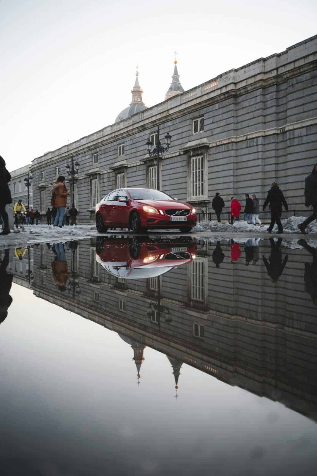 Auto rossa parcheggiata vicino a un edificio storico con riflesso sull'acqua, scena urbana con passanti, architettura antica e strade trafficate, in un contesto di città europea.