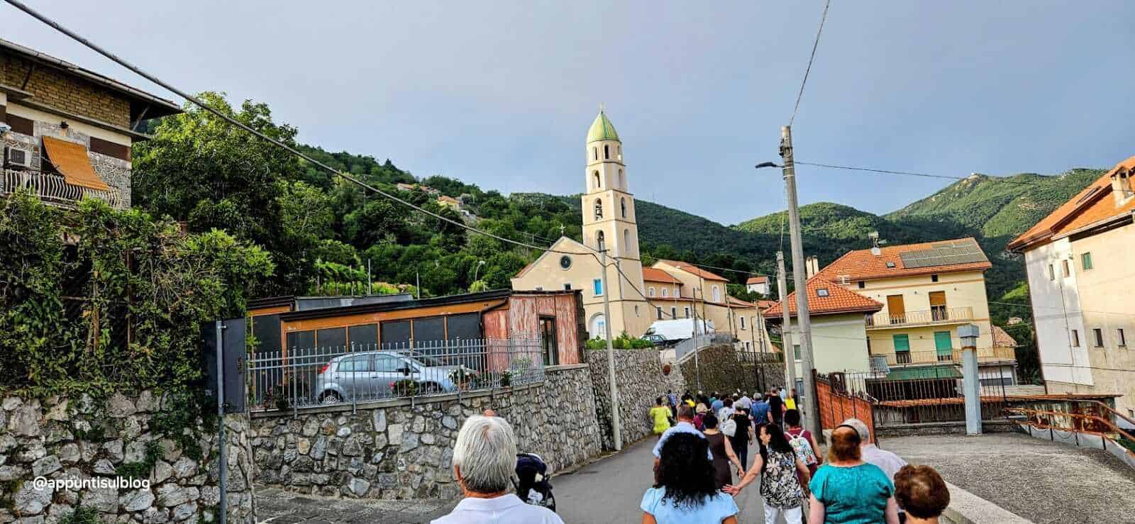 Cammino di turisti in un pittoresco villaggio italiano con chiesa storica, tra case con tetti in terracotta e paesaggi montani rigogliosi, perfetto per fotografie di viaggio, cultura e turismo in Italia.