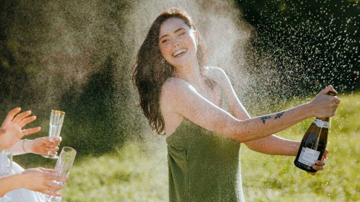 Festa all'aperto con donna felice che spruzza spumante durante un brindisi tra amici in un giardino soleggiato.